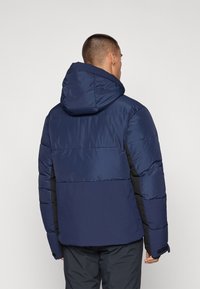 Navy blue puffer jacket with a hood, featuring quilted sections, black side accents, and textured fabric. Adjustable cuff straps included.