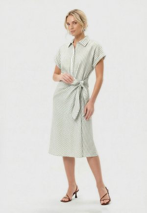 Kobieta w sukience midi w pionowe jasnoszare i białe paski, z krótkimi rękawami, kołnierzykiem, guzikami z przodu, paskiem w talii oraz brązowymi sandałami na obcasie.
