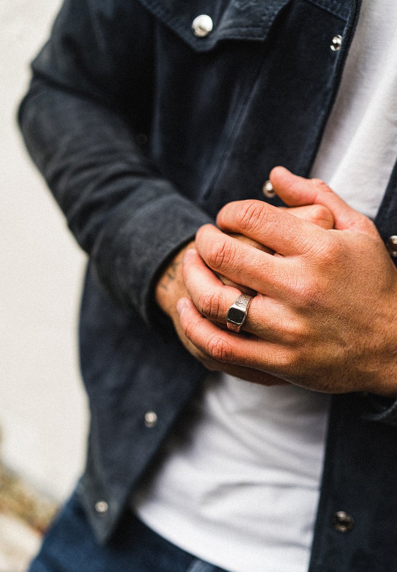 Vīrieša rokas saspiestas, tērptas tekstūratā melnā zamša jaku pār baltu t-kreklu. Uz kreisās rokas ir sudraba gredzens ar kvadrātveida melnu akmeni.