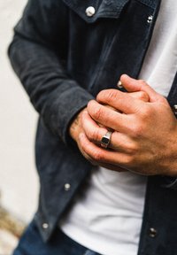 Rankos sujungtos kartu, ant piršto sidabrinis žiedas su juodu akmeniu, dėvint juodą švarką ir baltą marškinius.