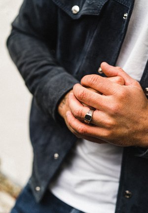 Mani intrecciate che indossano un anello d'argento con una pietra nera, vestite con una giacca nera e una camicia bianca.