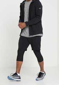 Outfit atletico composto da una giacca nera leggera, una maglietta grigia, pantaloni corti neri e leggings neri con sneakers a motivo in nero e blu.