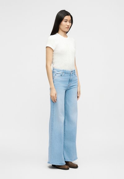 Νεαρή γυναίκα που στέκεται, φοράει λευκό τοπ με κοντά μανίκια, ανοιχτό μπλε παντελόνι με φαρδιές μπατζάκια και καφέ παπούτσια, μπροστά από ένα απλό φωτεινό φόντο.