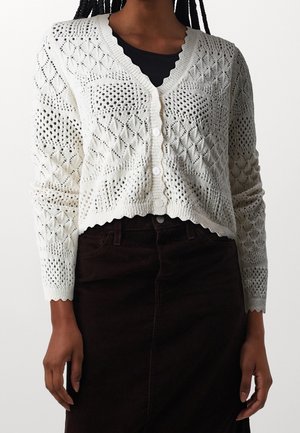 Bílý pletený cardigan s diamantovým vzorem, ve výstřihu do V a s vlnkovaným lemem. Obsahuje knoflíky po celé délce vpředu a dlouhé rukávy.