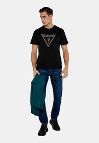 T-shirt di cotone nera con logo grigio triangolare, abbinata a jeans blu. Il modello tiene una felpa con cappuccio color teal. Sneakers nere completano l'outfit.