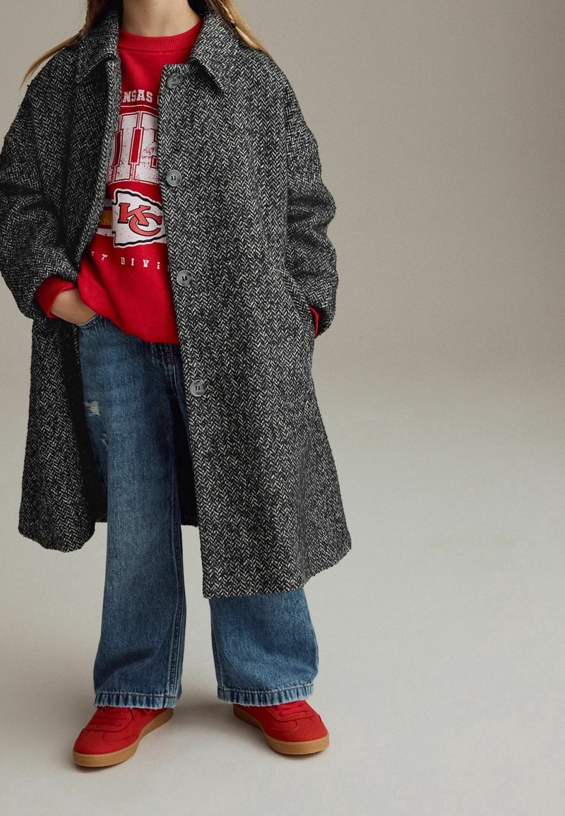 Personne portant un long manteau gris en chevrons sur un sweat-shirt rouge des Kansas City Chiefs, un jean large bleu et des baskets rouges, mains dans les poches.