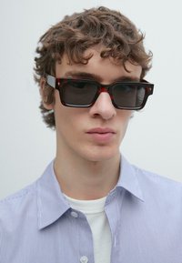 Occhiali da sole neri con aste lucide effetto tartaruga e lenti scure. Il modello indossa una camicia a righe azzurre sopra una maglietta bianca.