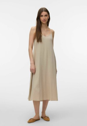 Jeune femme aux longs cheveux bruns portant une robe midi beige sans manches et des chaussures plates marron, debout contre un fond blanc.