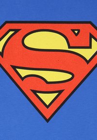 Červený a žlutý odznak štítu Superman na modrém pozadí, zobrazující stylizované písmeno "S" uvnitř diamantového tvaru.