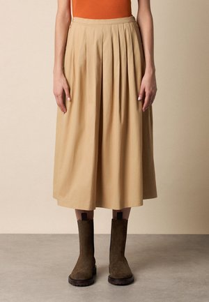 Kobieta ubrana w beżową midi spódnicę z wysokim stanem i plisami z przodu, w połączeniu z ciemnobrązowymi zamszowymi botkami, stoi na szarej podłodze.