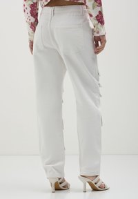 Frau trägt weiße High-Waist-Hosen mit Gesäßtaschen und weiße Absatzsandalen, von hinten vor einem schlichten Hintergrund gezeigt.