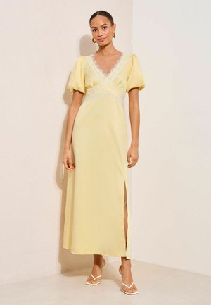 Frau steht in einem pastellgelben Midikleid mit Puffärmeln, V-Ausschnitt mit Spitzenbesatz und seitlichem Schlitz, trägt weiße Sandaletten mit Absatz.