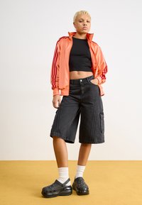 Koralle Satinjacke mit roten Streifen, schwarzem Cropped-Top, schwarzen Denim-Cargo-Shorts und schwarzen Clogs, kombiniert mit weißen Socken auf einem gelben Hintergrund.