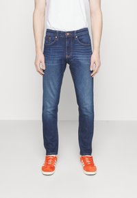 Mörkblå jeans i denim med smal passform, utrustade med fem fickor, knappstängning och subtila blekningsdetaljer. Matchade med ljusorange sneakers.