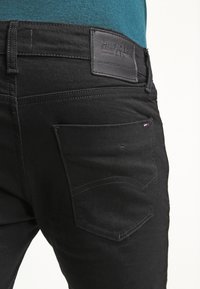 Černé denimové džíny s koženou nášivkou na pasu, jednoduchými švy a rovnými kapsami.