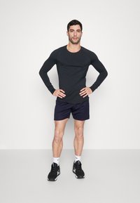 Crna dugih rukava sportska majica s raglan rukavima, u kombinaciji s tamnoplavim šortsevima. Model nosi crne sportske tenisice s bijelim detaljima.