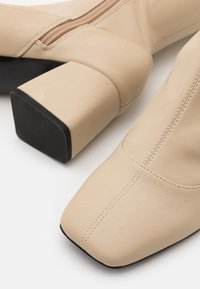 Beige keinonahkaiset nilkkurit, joissa on neliömäinen kärki, teksturoitu pinta ja jykevä musta korko. Varustettu sivuvetoketjulla kiinnittämistä varten.