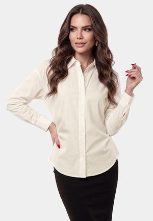 Donna con lunghi capelli mossi castani che indossa una camicia crema con bottoni e gonna nera, in posa con una mano sul fianco e l'altra sollevata.