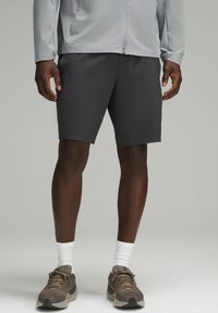 Pantaloni corti da uomo grigio scuro, tessuto morbido, fino al ginocchio, abbinati a una giacca leggera grigio chiaro con zip, scarpe da ginnastica marroni e calze bianche.