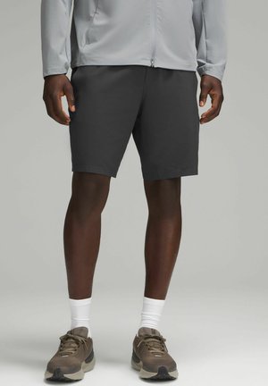 Pantaloni corti da uomo grigio scuro, tessuto morbido, fino al ginocchio, abbinati a una giacca leggera grigio chiaro con zip, scarpe da ginnastica marroni e calze bianche.