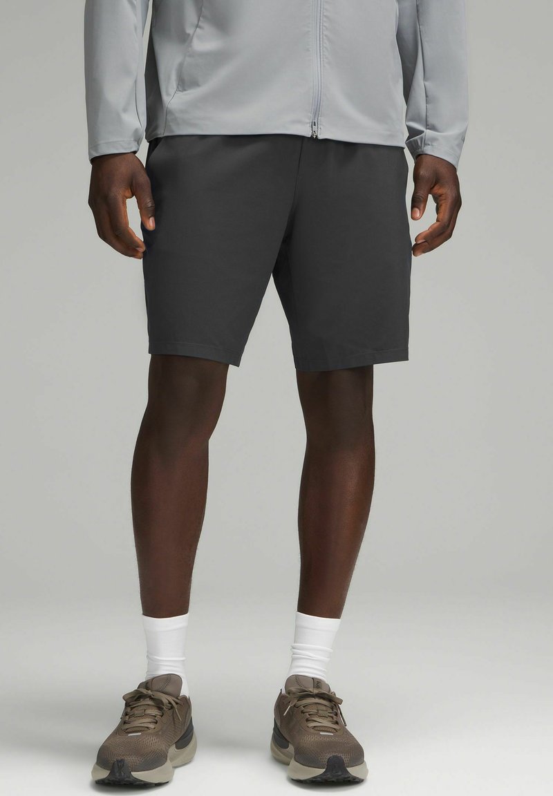 Pantaloni corti da uomo grigio scuro, tessuto morbido, fino al ginocchio, abbinati a una giacca leggera grigio chiaro con zip, scarpe da ginnastica marroni e calze bianche.