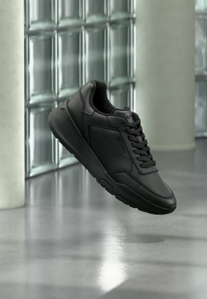 Baskets en cuir noir avec lacets flottant au-dessus d'un sol gris réfléchissant devant un mur en blocs de verre.