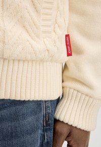 Pull en tricot crème à motif torsadé, avec un ourlet et des poignets côtelés. Comprend une étiquette rouge avec "JOOP! JEANS." Assorti à un jean en denim bleu.