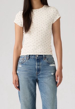 Femme portant un haut blanc à manches courtes avec de petits imprimés floraux et un jean bleu taille haute avec une légère usure.