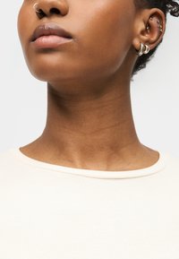 Κοντινό πλάνο του λαιμού και του προσώπου ενός ατόμου, που φοράει ασημένιο κρίκο στη μύτη και πολλαπλές τρυπητές στα αυτιά, με ανοιχτόχρωμο ριμπ πλεκτό τοπ.