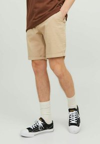 Shorts beiges avec des ourlets roulés, en tissu léger. Associés à des baskets noires et des chaussettes blanches. Haut brun partiellement visible.