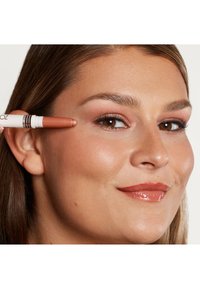 Crayon de maquillage pour les yeux marron et blanc tenu près de l'œil, avec une finition lisse et mate. Le modèle montre un gloss à lèvres aux tons chauds et un fard à paupières subtil.