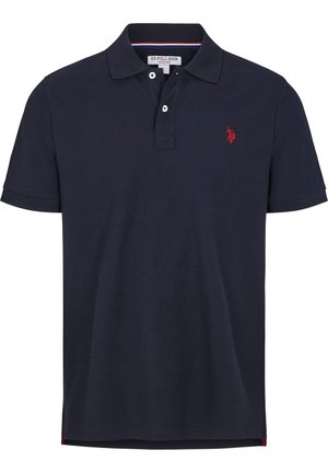 Mand iført en marineblå polo med korte ærmer med rød broderet logo og beige bukser, stående mod en ensfarvet baggrund.