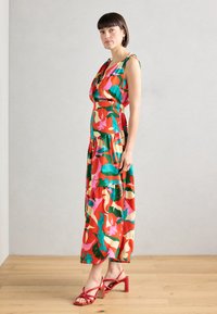 Robe maxi colorée avec un motif floral en rouge, rose, vert et crème ; décolleté en V et design à niveaux ; associée à des talons strass rouges.