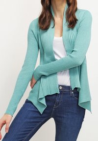 Cárdigan verde menta acanalado con un frente abierto, mangas largas y un dobladillo asimétrico, combinado con una blusa blanca y jeans azul oscuro.