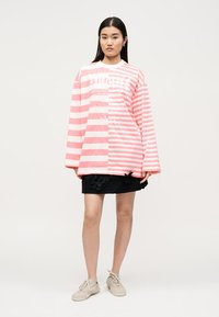 Femme debout portant une chemise oversize à manches longues rayée rose et blanche, une jupe noire avec des accents floraux et des baskets beiges sur un fond uni.
