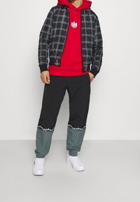 Zwart-wit geruite bomberjack over een rode hoodie, gecombineerd met zwarte en turquoise broek met een bloemenpatroon aan de zoom. Witte sneakers.