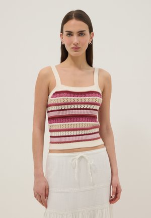 Femme aux cheveux raides et foncés portant un haut en maille sans manches avec des rayures roses et crème et une jupe blanche à cordon sur un fond uni.