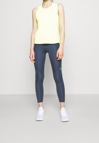 Sportovní tílko v neonově žluté barvě, spárované s námořnickými legínami s texturovaným vzorem a bílými sportovními botami.