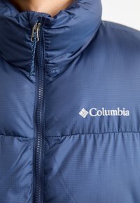 Námořnická prošívaná bunda s vysokým límcem, předním zipem a texturovanou látkou s jemným proužkovaným vzorem. Logo Columbia na hrudi.