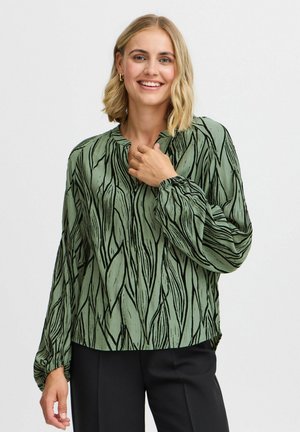 Green blouse with black leaf patterns, long sleeves, and a loose fit. Features a round neckline and gathered cuffs. Smooth texture.