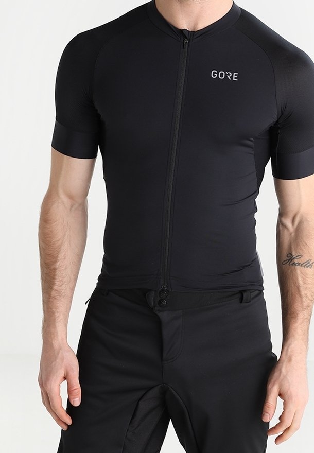 Jersey de ciclismo negro de manga corta con cremallera frontal completa, textura suave y logo en la parte superior izquierda. Combinado con pantalones cortos de ciclismo negros.