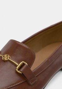 Mocassin en cuir brun avec un ornement décoratif en métal doré sur la bride, présentant une texture lisse et un design à bout arrondi.