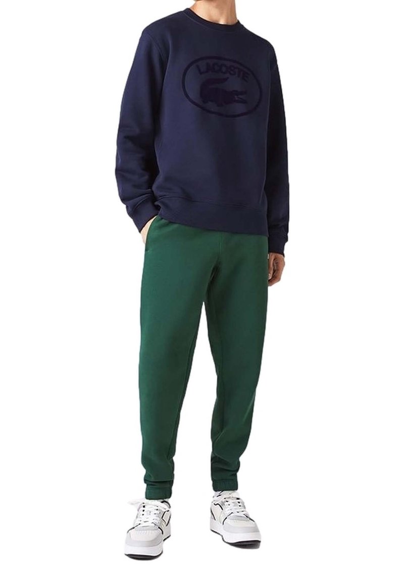 Laivaston sininen huppari, jossa on kirjailtu Lacosten logo, yhdistettynä vihreisiin collegehousuihin ja valkoisiin tennareihin. Materiaalit näyttävät pehmeiltä ja rentoilta.