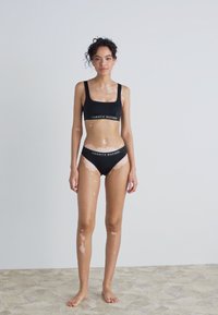 Črni športni nedrček in uskladen bikini z belimi podrobnostmi logotipa. Gladek material, minimalističen dizajn in prilegajoč videz. Svetla koža z madeži.