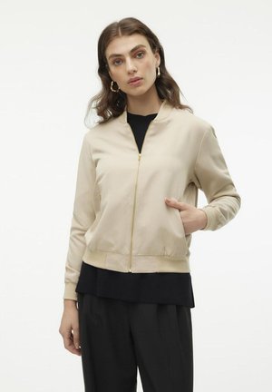 Beige bomber jacket with a front zipper, ribbed cuffs, and hem. The model wears a black shirt underneath, with a side pocket on the jacket.