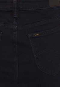 Jeans de denim azul oscuro con etiqueta "Lee" dorada en contraste, diseño de cinco bolsillos, costuras visibles y una textura suave.