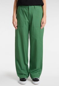 Groene cargobroek met wijde pijpen, gemaakt van zachte stof, met twee grote voorzakken en een gladde textuur. Draagt met zwarte sneakers.