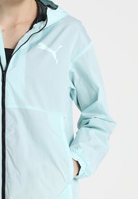 Ljust blå vindjacka med dragkedja framtill, elastiska ärmslut och Puma-logotyp i vitt. Tillverkad av lätt, texturerat tyg.