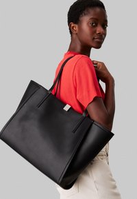 Sac fourre-tout en cuir noir avec deux longues poignées, base plate et une petite décoration en argent. Le sac est tenu contre un modèle portant une chemise rouge.