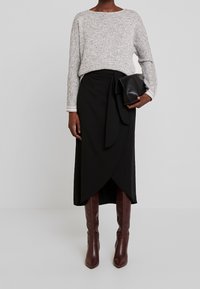 Person, die einen hellgrauen Strickpullover, einen schwarzen Wickelrock und braune Stiefel bis zu den Knien trägt, hält eine schwarze Ledertasche vor einem schlichten Hintergrund.